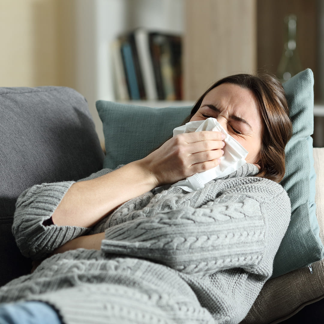Donna sul divano che starnutisce con un fazzoletto — sintomi da Virus Respiratorio Sinciziale, infezione delle vie respiratorie negli adulti